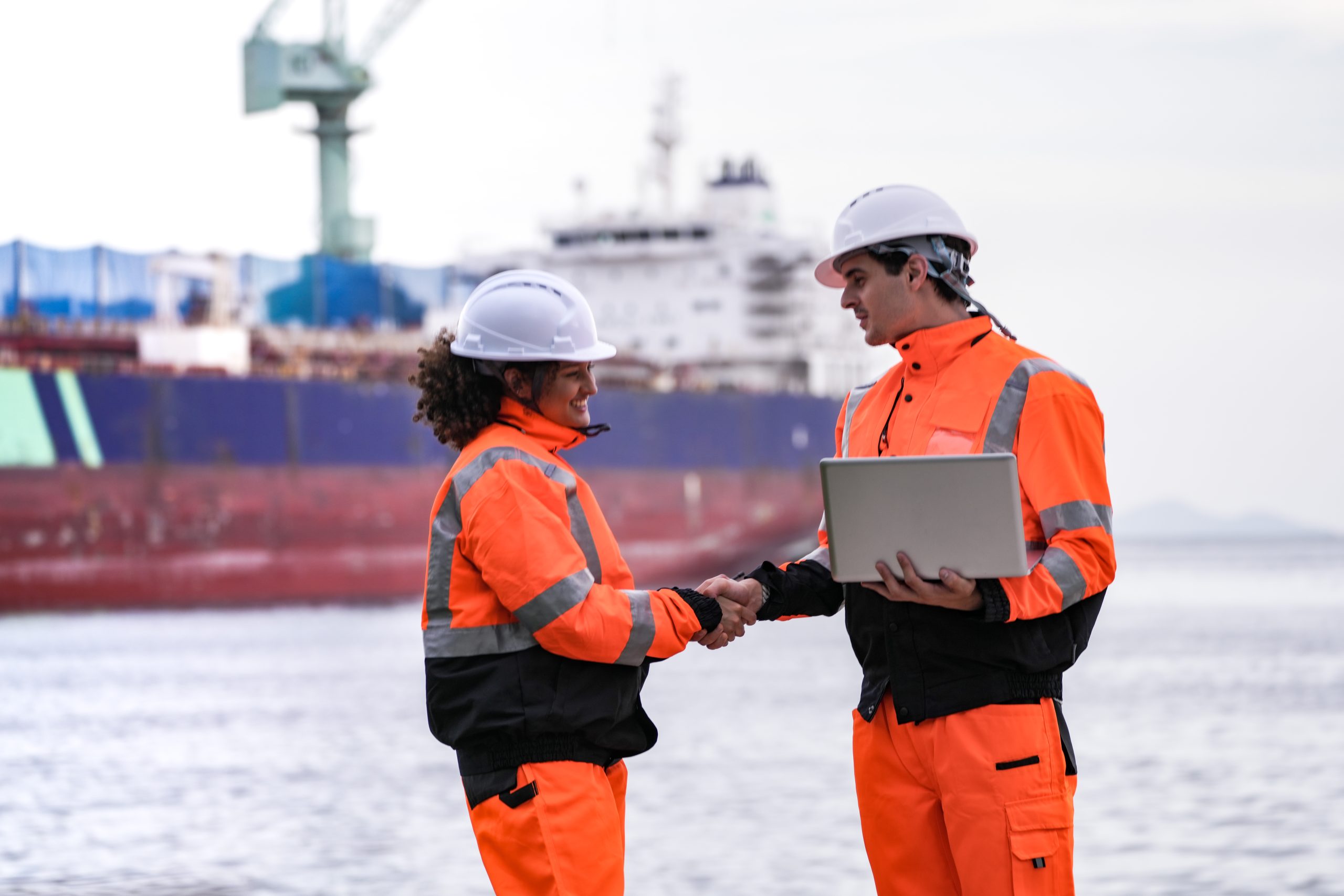 Two port engineers in high-visibility orange uniforms and hard hats communicate while inspecting dockside logistics using a laptop and walkie-talkie, symbolizing teamwork and safety.