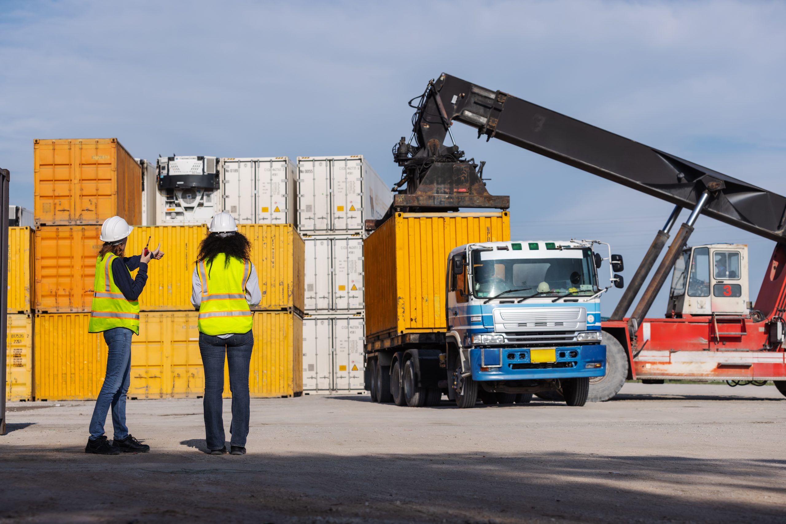 Two female workers in safety vests and hard hats oversee container loading operations at a shipping yard. A crane lifts a yellow container onto a truck, highlighting teamwork and global logistics.